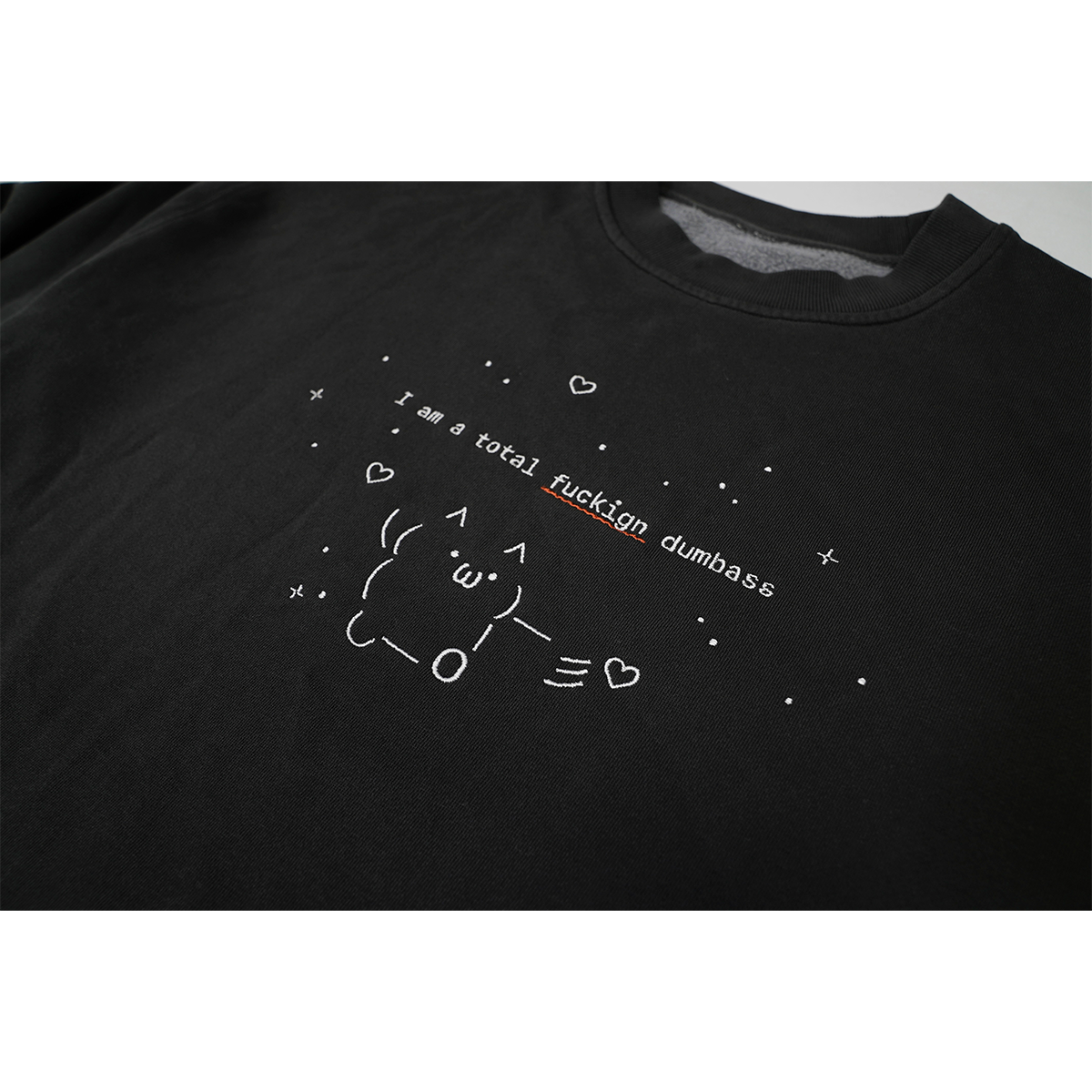 Detailed close-up of the white print across the chest reading “I’m a total fuckign dumbass,” with a red squiggly underline beneath the misspelled word “fuckign.” The text is surrounded by a small figure and fine line illustrations of stars and hearts.