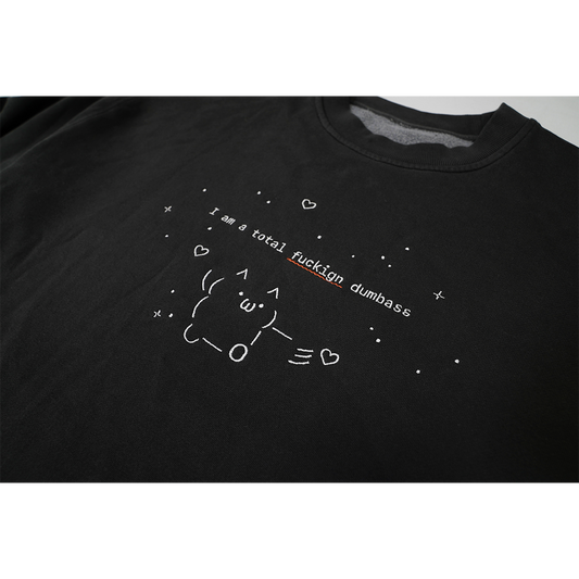 Detailed close-up of the white print across the chest reading “I’m a total fuckign dumbass,” with a red squiggly underline beneath the misspelled word “fuckign.” The text is surrounded by a small figure and fine line illustrations of stars and hearts.